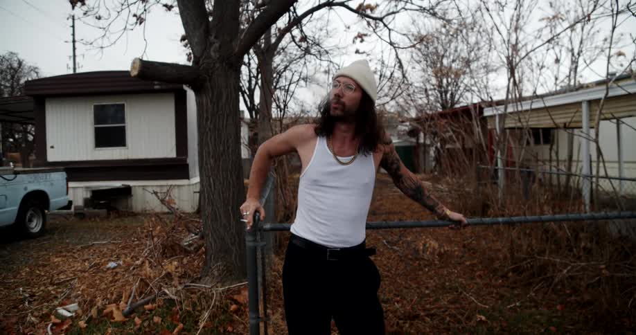 A man with glasses smoking a cigarette with houses trees the sky and clouds in the background