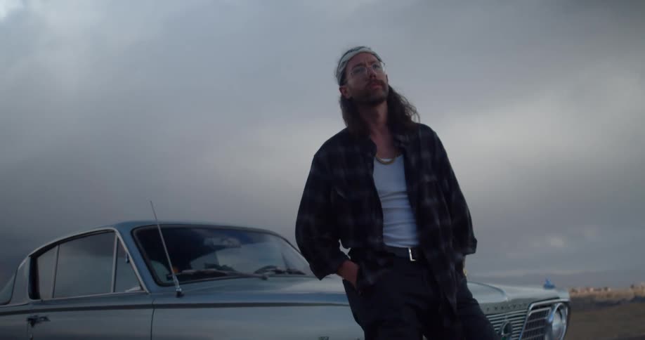 A man stands outside looking up at the cloudy sky with a vintage car behind him