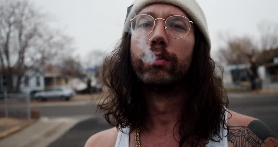 A man with glasses smoking a cigarette with houses trees the sky and clouds in the background