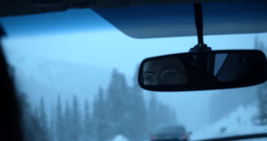A driver runs his vehicles windshield wipers as he follows another car down a narrow road through a snow covered forest