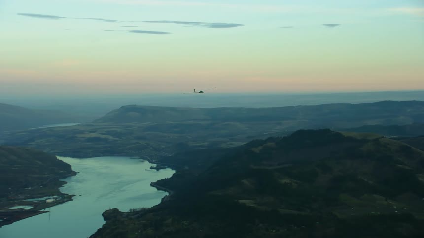Air to air shot of a small white glider airplane gliding over a river and then turning right towards camera revealing the Columbia River before disappearing out of frame in this air to air aerial at sunset
