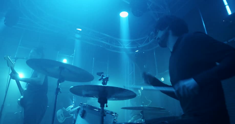 A man drums as his band plays on stage under blue lights
