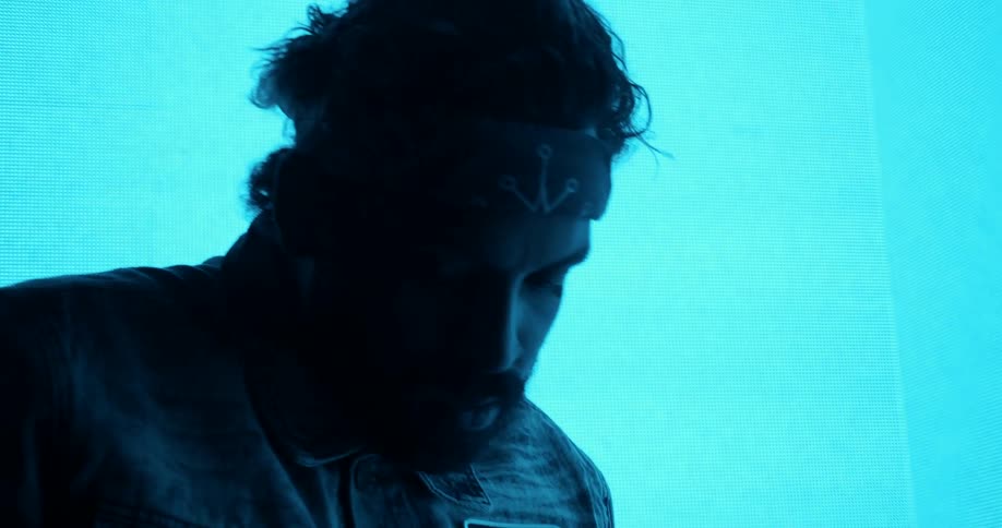 A man in a bandana and denim jacket poses in front of a large screen with flashing colorful shapes