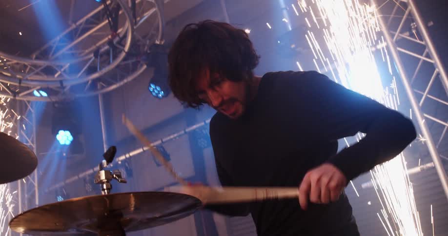 A man sitting on stage playing drums with drumsticks with fireworks and lights in the background