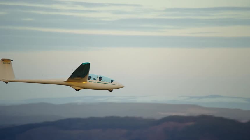 The Camera passes a white no engine glider where the pilot and the passenger are briefly visible before banking a hard left turn and leaving the frame in this air to air aerial shot