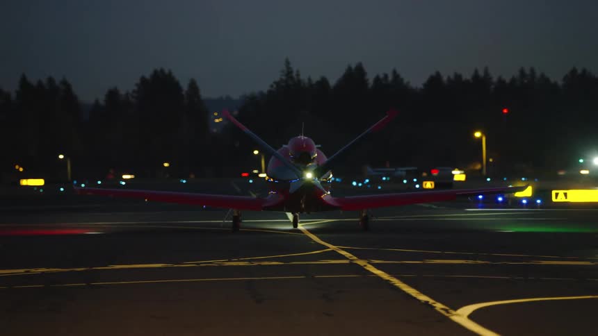 A post sunset night shot of a v-tail Beechcraft Bonanza taxiing at an airport at night with a white strobe light flashing and yellow taxi way paint lines on the tarmac