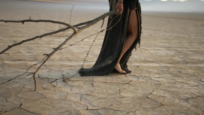 A woman drags a long branch and barbed wire behind her as she walks barefoot through the desert
