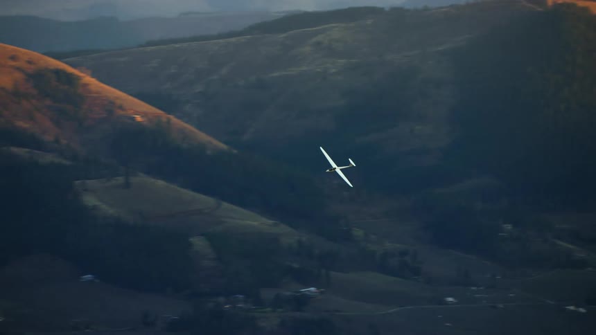 A white no engine glider aircraft makes a sweeping left hand turn with hills illuminated by golden light and then revealing the Columbia River Gorge