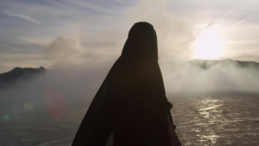 Woman wearing a hooded cloak is walking across a dry desert with a bright sun and mountains in the distance