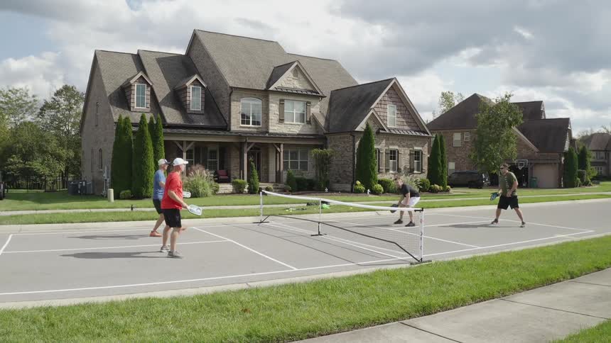 Friends playing pickleball on a court in front of a large house in a neighborhood