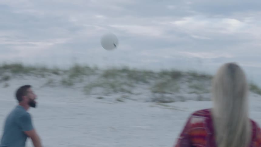 A man and two women on a beach playing with a ball with the sky and clouds in the background
