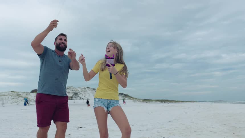 A father and daughter laugh on the beach as they try and reel a kite in