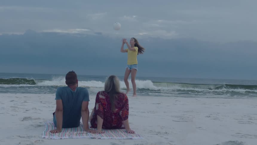 Father and mother watching their teenage daugter playing with a volleyball on the white sand of a beach near the ocean