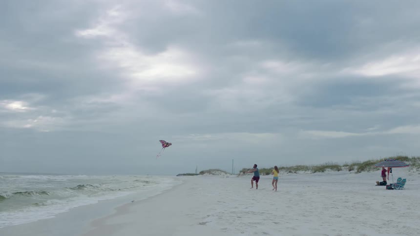 A man and a woman in a beach flying a kite with the sky and clouds in the background