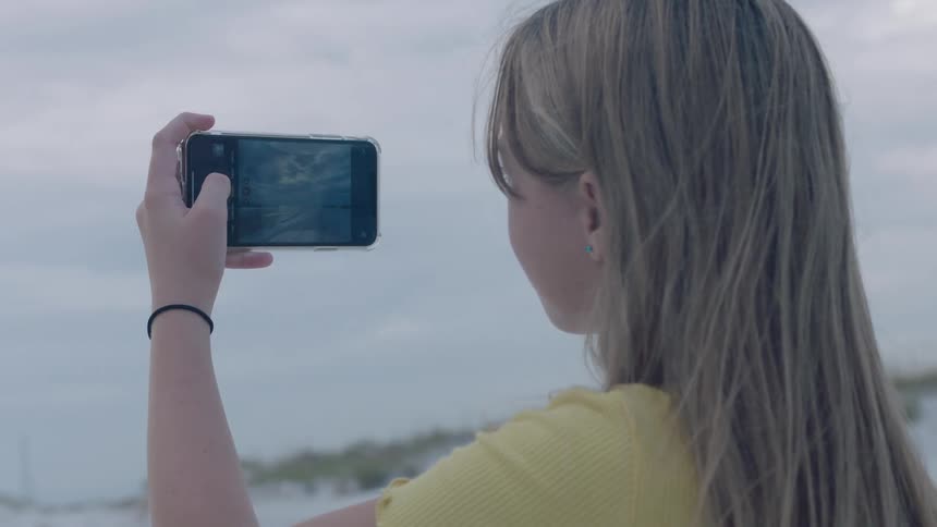 Teenage girl standing on a beach filming the ocean with her smartphone