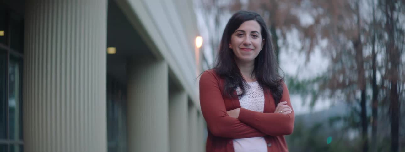 Woman with dark wavy hair stands arms crossed and smiles at the camera next to building