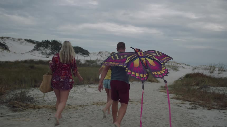 Two women and a man with a kite walking on a beach with the sky and clouds in the background