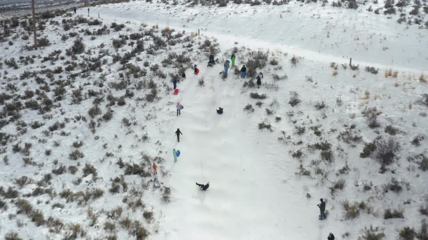 People sliding down a snowy slope on sleds with other people standing and walking