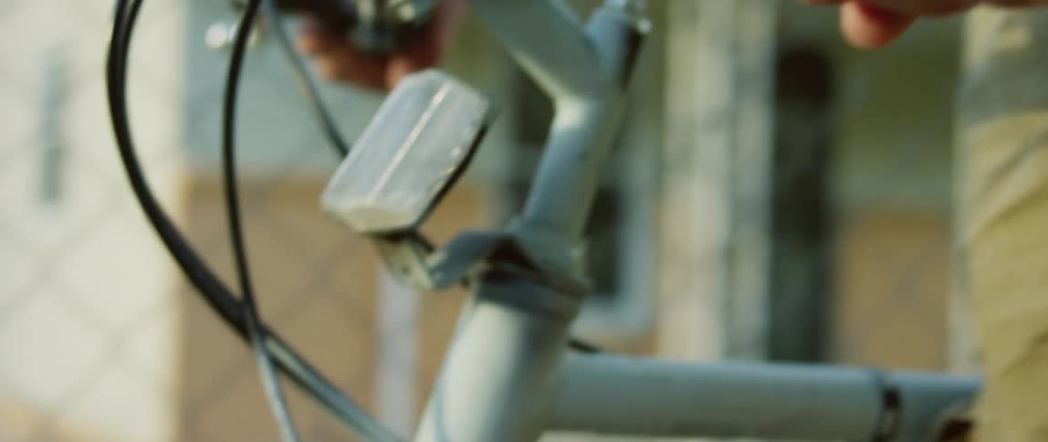 Hands hold the handlebar and guide the bike as a person walks past a fence and home