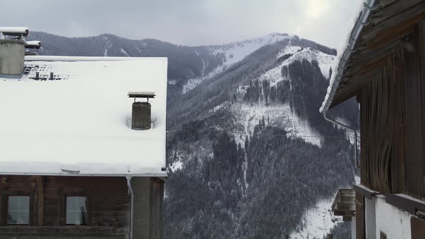 A couple of tall wooden buildings bordering snowy mountains covered in forest on a snowy day