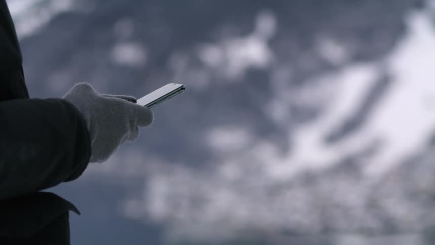 A person with gloves using a smart phone with a mountain range and snow in the background