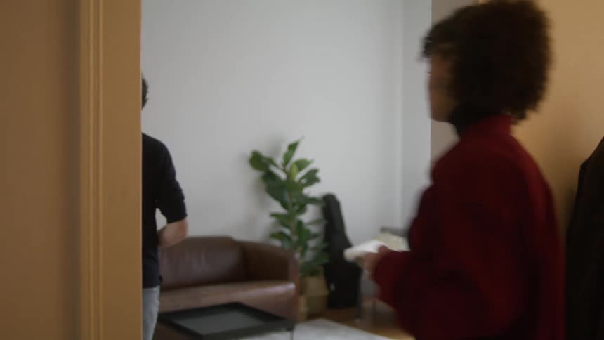 A man drinking a beer stands in a hallway at home and talks to a woman and then they walk in to another room and he sits on a couch