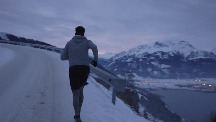 A man runs along a snow covered highway above the shore with mountains in the background