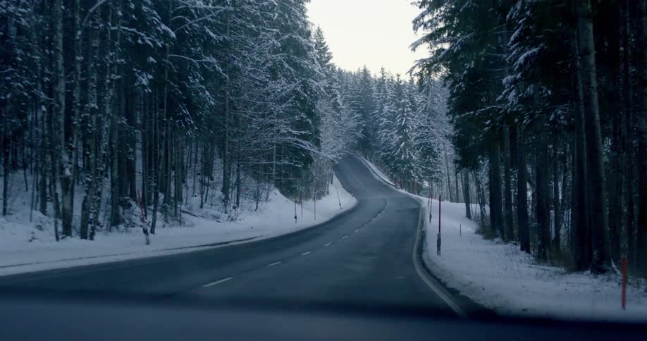 A vehicle with a windshield driving on a road surrounded by snow and trees with the sky and clouds in the background