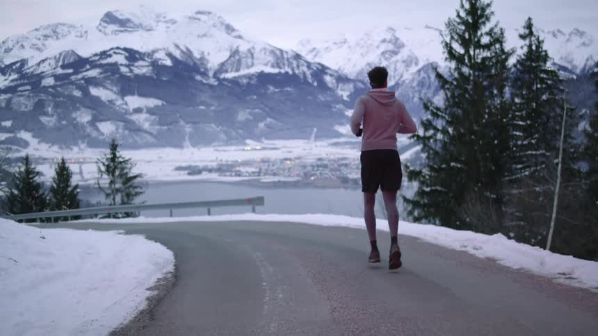 A man jogging on a street with a lake a mountain range the sky and clouds in the background