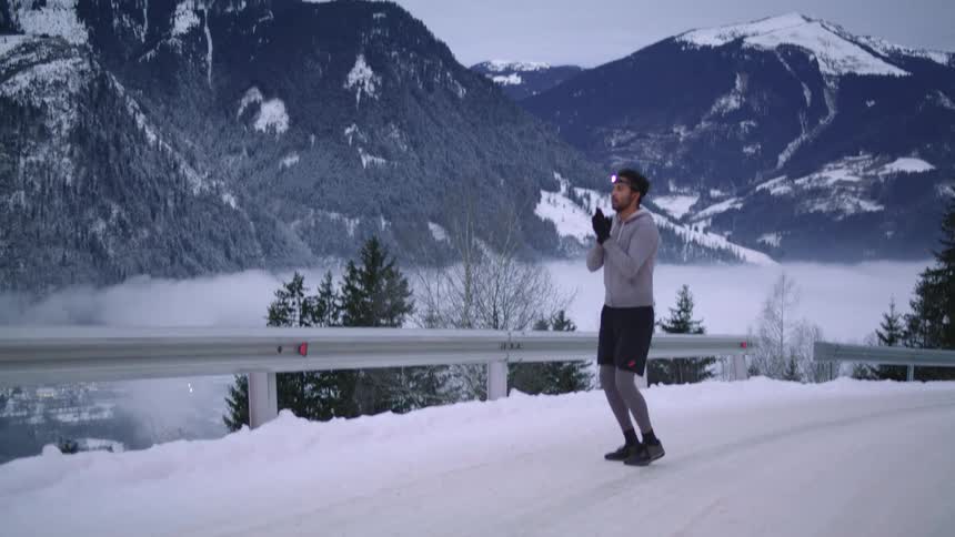 A man with a flashlight on his head with a hoodie and gloves jogging on a road with a mountain range the sky and clouds in the background