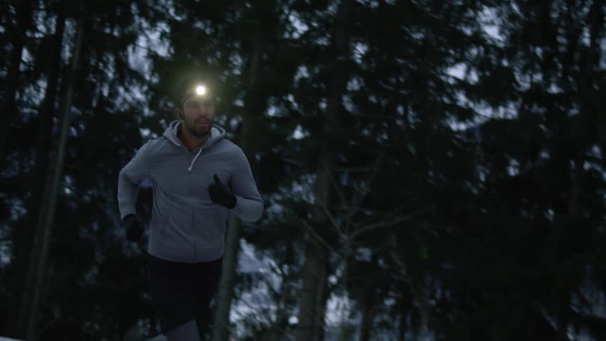 A man with a headlight on running outdoors on a road in a forested area that is covered in light snow