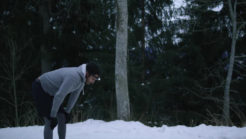 A man appears to be taking a break from running as he bends over to catch his breath and then pulls his hood on