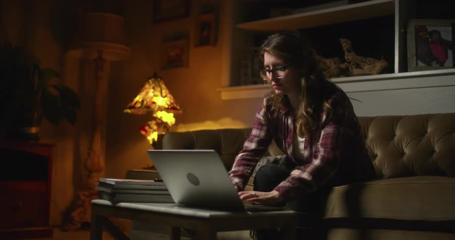 Woman sitting on a brown couch in a living room at night opens up a laptop computer in front of her on a coffee table and starts typing