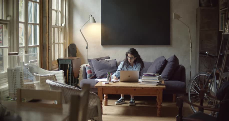 A young woman sits on a couch at home and looks at a laptop as she writes