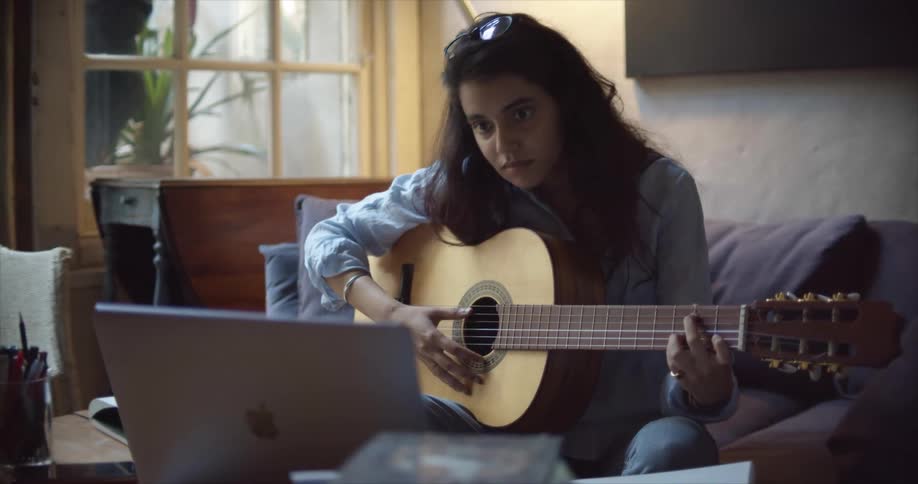 A female sits on a sofa while playing a guitar and looking at the screen of a laptop