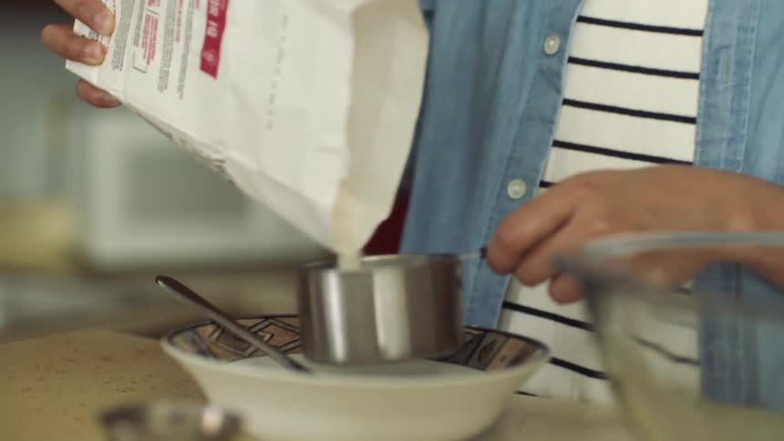 A person pouring flour into a metal measuring cup in a home kitchen