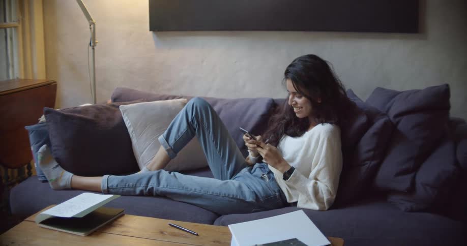 A woman sitting on a small couch in a living room smiling and taking selfies with a smart phone