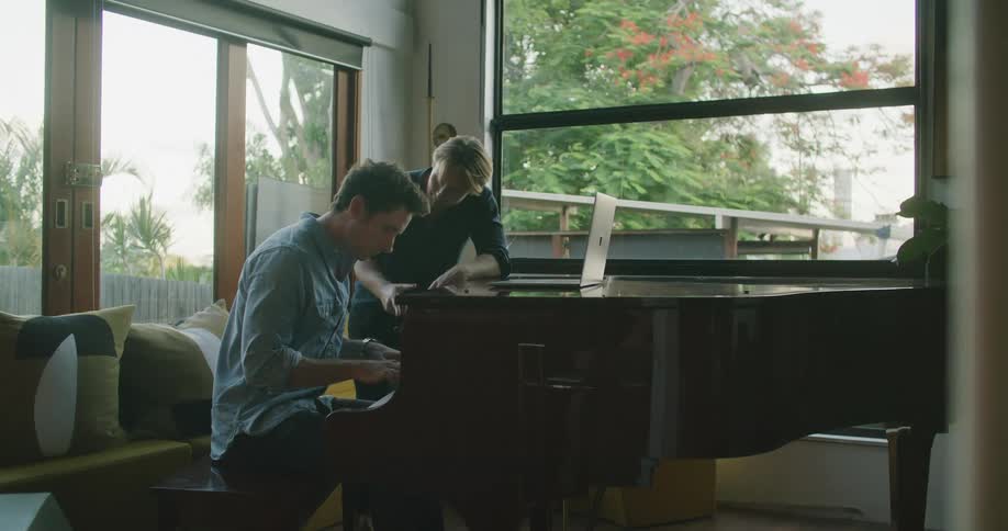 Two men in a room as one plays the piano with a laptop on top of it with a window in the background