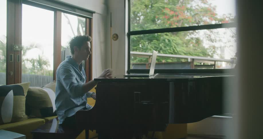 A man in a room sitting and playing a piano while watching a laptop with a window in the background