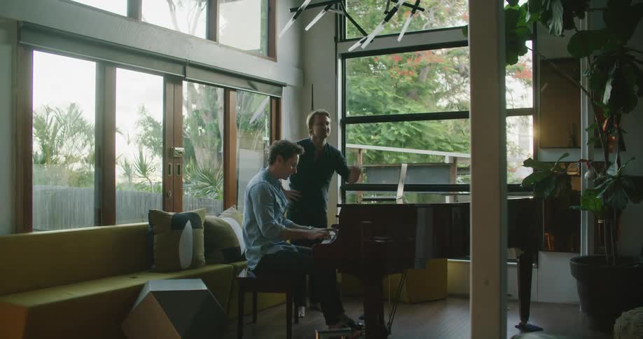 Two men in a room as one plays the piano with a laptop on top of it with a window in the background