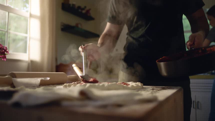 A person in the kitchen adding tomato sauce on top of pizza dough on a counter