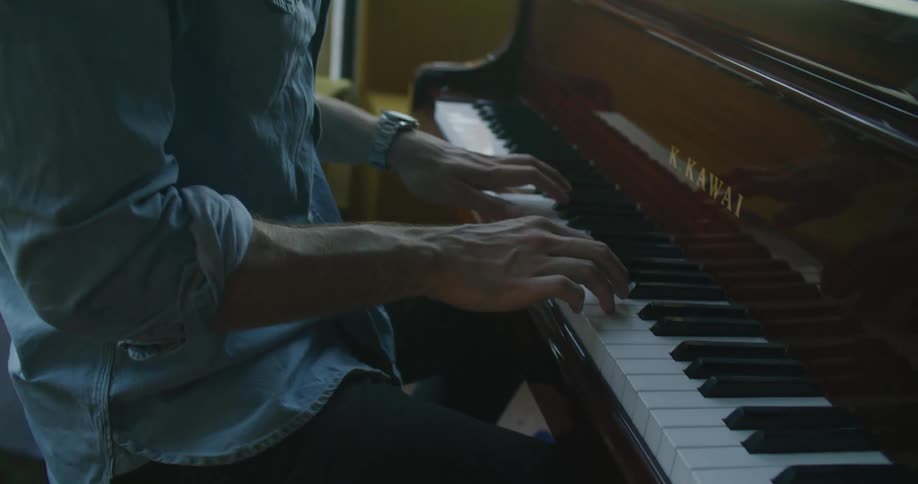 A man in a room playing the piano with a laptop on top with a window in the background
