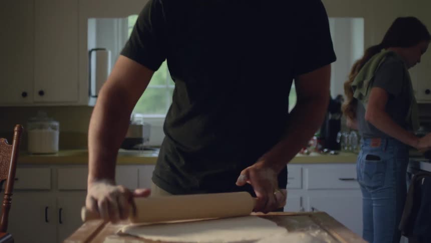 A man cooking in a kitchen at home with a woman uses a rolling pin to roll dough out flat on a countertop at daytime