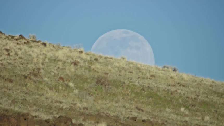 Aerial looking up at a mostly full moon in the afternoon blue sky before a rocky and grass covered ridge wipes the sky from view