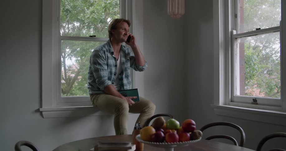 A man smiles as he talks on the phone while sitting on the window sill then looks bored after hanging up
