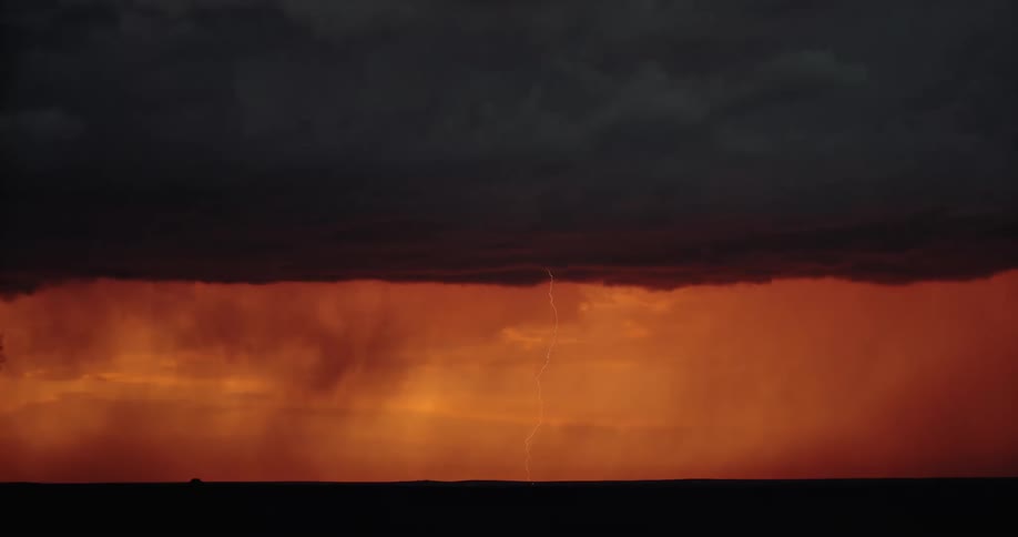 Lightning strikes straight down into a field in the distance from dark storm clouds in an orange sky at sunset