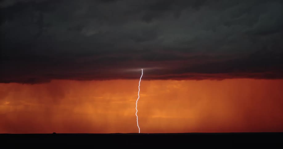 Lightning strikes from a sky of thick storm clouds illuminate a flat countryside under an orange sky