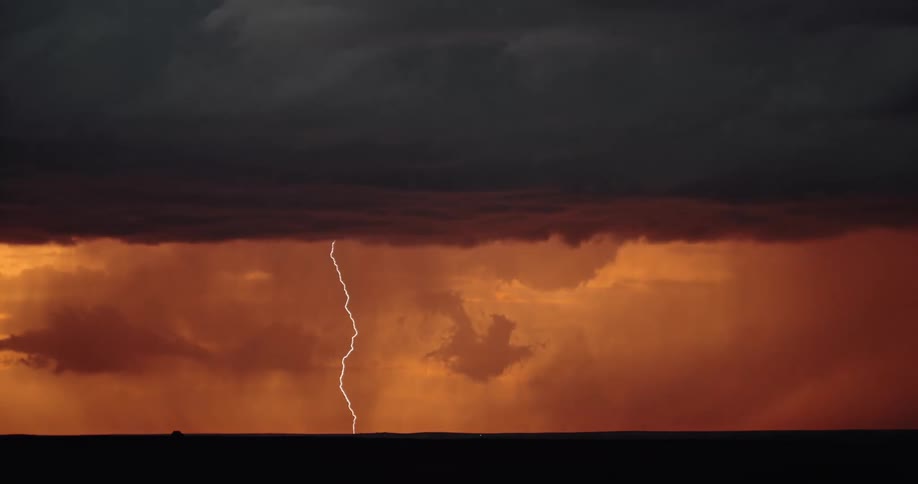 Lightning strikes down into a field from dark storm clouds in an orange glowing sky