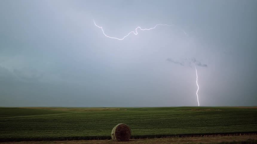 Lighting strikes across storm clouds in the sky and down onto the ground in the distance of a field at daytime