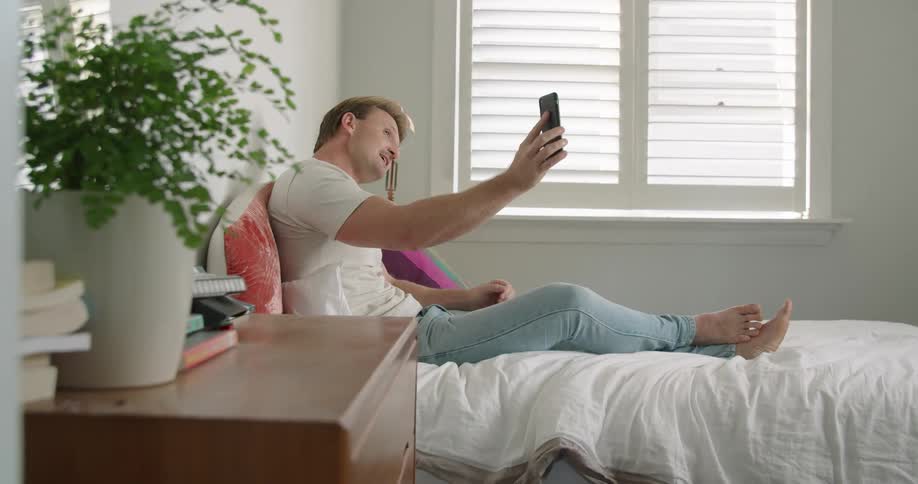 A man sits on a bed in a bedroom while video chatting on a smartphone at daytime
